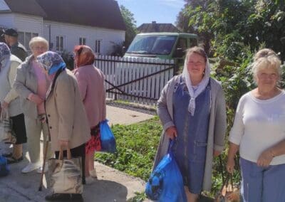 A group of women, most wearing headscarves, stand outdoors holding blue plastic bags as part of Food Aid for Northern Ukraine, with houses, a fence, and greenery in the background.