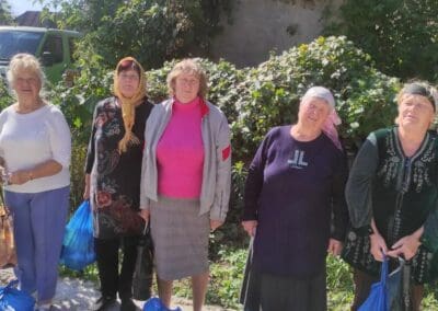 A group of seven elderly women stand outdoors on a sunny day, each holding blue bags provided by Food Aid for Northern Ukraine, with greenery and a parked vehicle in the background.