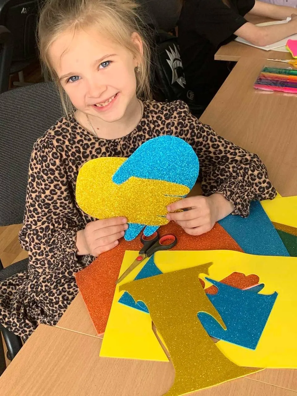 A young girl holds glittery cutouts shaped like hands, sitting at a table with colorful craft materials and smiling at the camera.