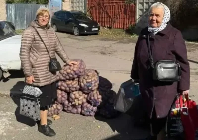 Two older women stand on a street near several large bags of potatoes, each holding shopping bags. A white car and fence are in the background.