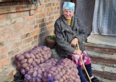 An elderly woman with a cane sits beside several large bags of potatoes in front of a brick building and steps.