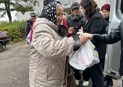 An elderly woman in a light coat receives a plastic bag of supplies from a volunteer while others wait in line outside a building.