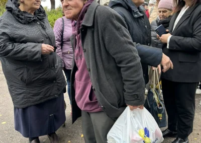 A group of elderly people stand outdoors in autumn clothing, some holding bags, next to a woman in formal attire, in front of a gray building.
