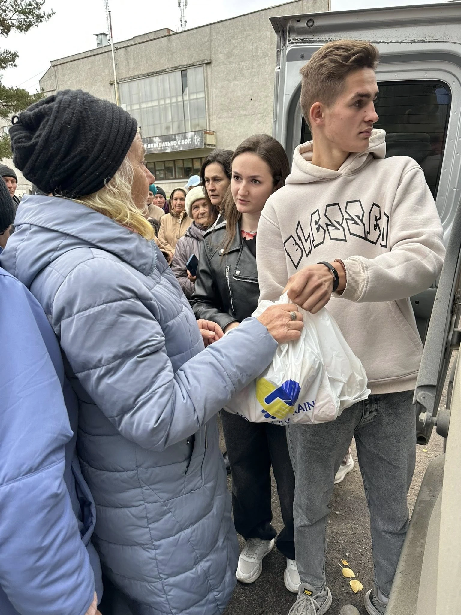 People receive aid bags labeled "Ukraine" from a vehicle while standing in a crowded outdoor line.