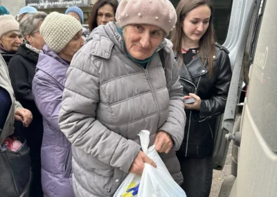 An elderly woman in a grey coat and pink hat holds plastic bags while standing near a van, surrounded by other people in winter clothing.