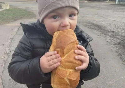 A young child in a jacket and beanie stands outdoors, holding and biting into a large loaf of bread.