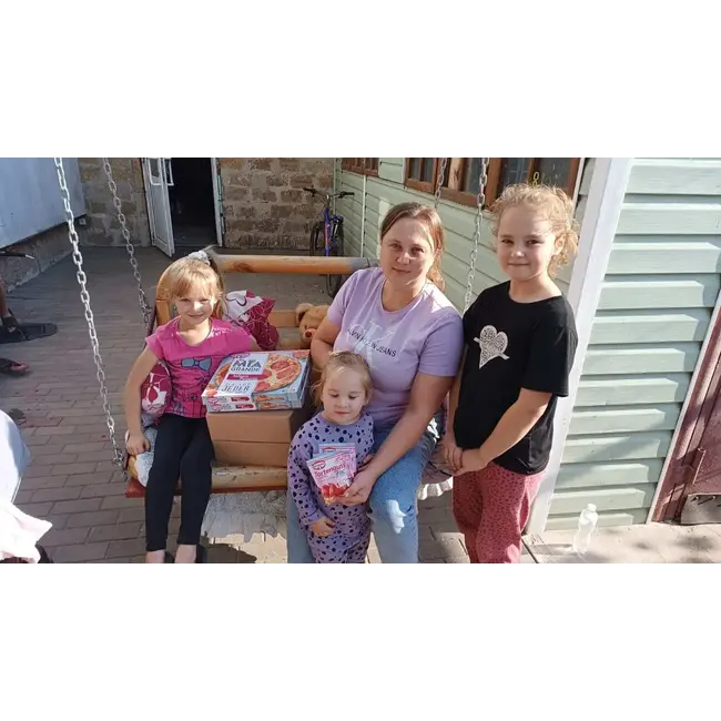 A woman and three young girls sit and stand around a wooden swing bench outdoors, with a box of food items visible beside them.