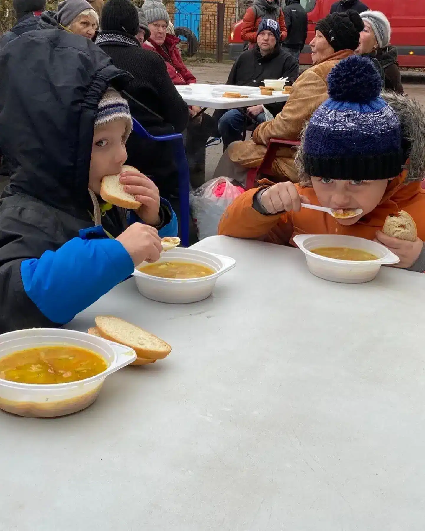 Two children in winter clothing eat soup and bread at an outdoor table, with several adults sitting in the background.