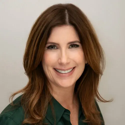Smiling woman with long brown hair wearing a dark green shirt, posing in front of a plain light background.