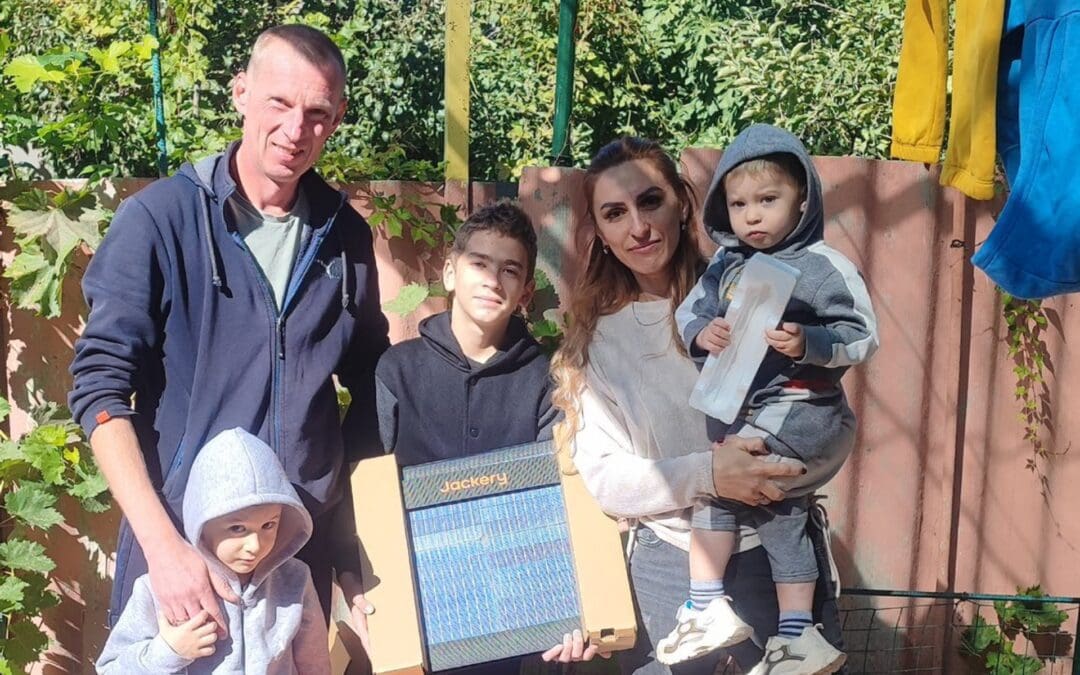 A family of five poses outdoors, one child holding a Jackery solar panel box—symbolizing Hope For Ukraine humanitarian impact. Dressed in casual clothing and hoodies, they stand together with shrubs and trees in the background.