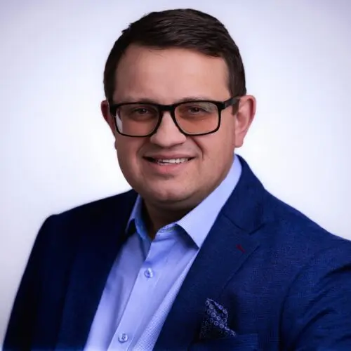 Man wearing glasses, a blue blazer, and a light blue shirt, smiling at the camera against a plain, light background.
