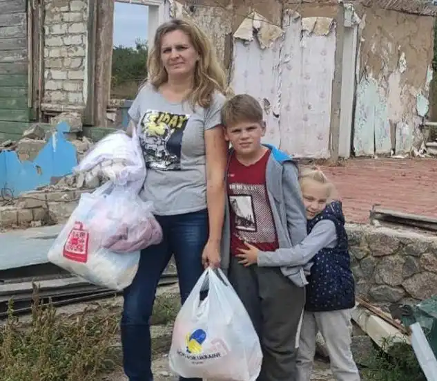 A woman and two children stand in front of a damaged building, holding bags of belongings. The children appear close to the woman, and the scene suggests displacement or hardship.