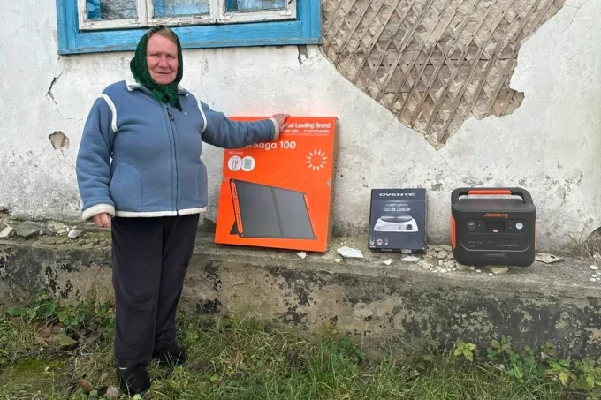 A person in blue winter coat stands outdoors holding a portable Jackery solar panel by a rural road.