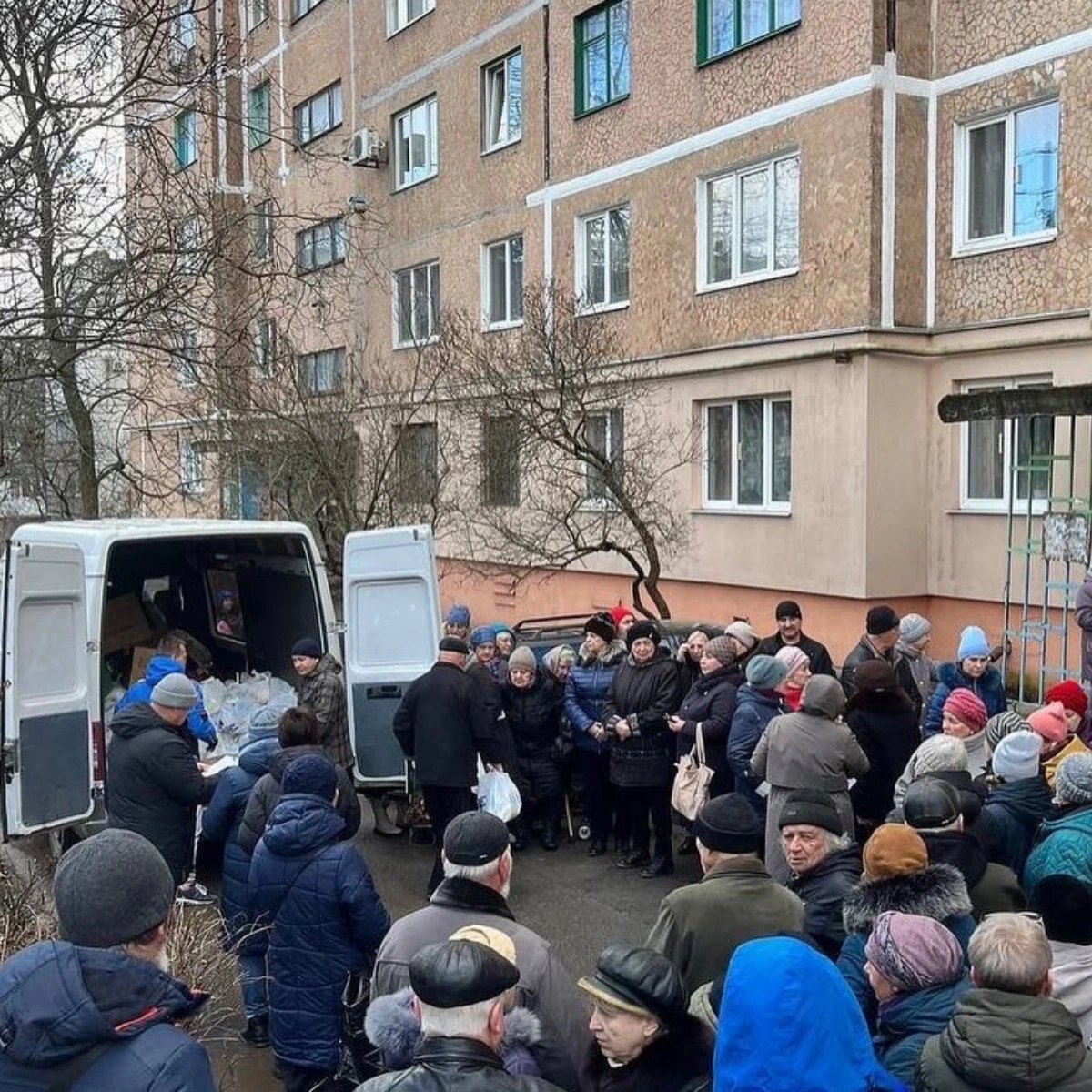 A crowd of people stands in line outside a van distributing supplies in front of a multi-story apartment building.