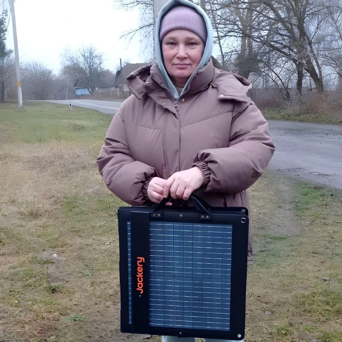 A person in a brown winter coat and pink beanie stands outdoors holding a portable Jackery solar panel by a rural road.