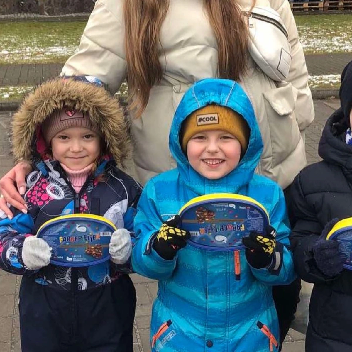 Three children in winter jackets and hats stand outside, smiling and holding oval blue tins; an adult stands behind them with a hand on one child's shoulder.