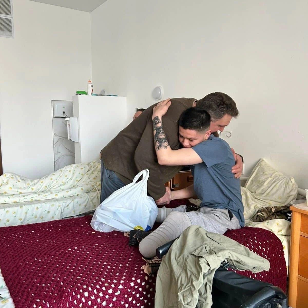 Two people embrace on a bed in a simply furnished room. One has a prosthetic leg and sits while hugging the other, who stands and leans over. A plastic bag rests on the bed.