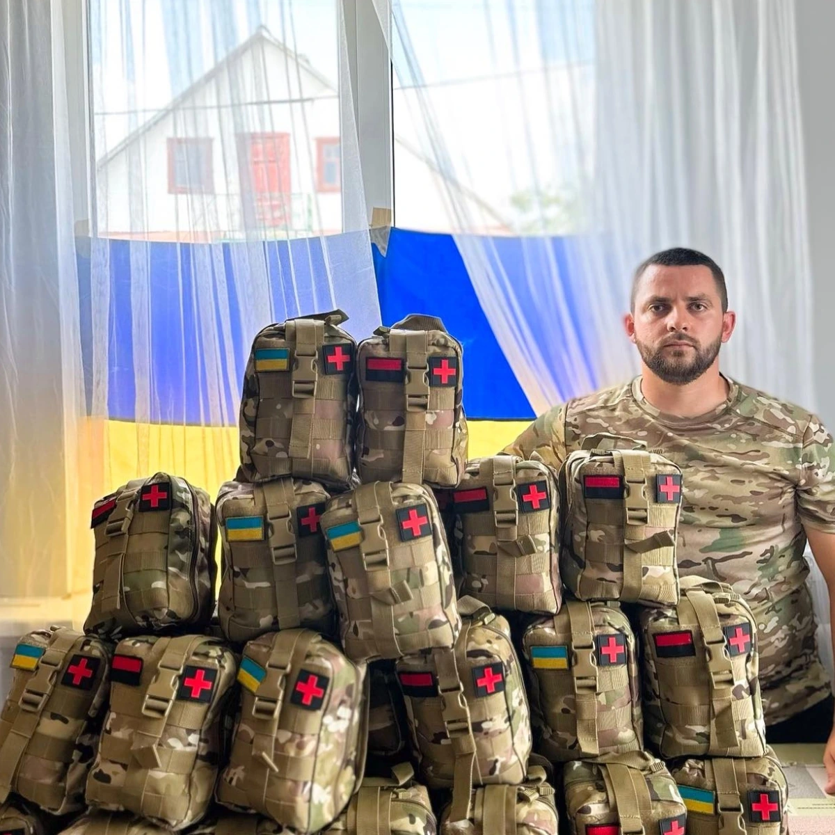 A man in camouflage sits beside a stack of military medical pouches with red cross symbols; a Ukrainian flag hangs in the background.