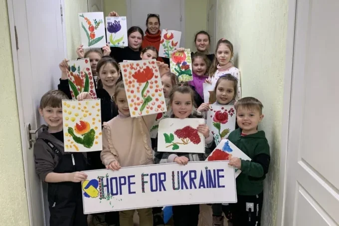 A group of children stand in a hallway, holding up colorful artwork and a sign that reads 