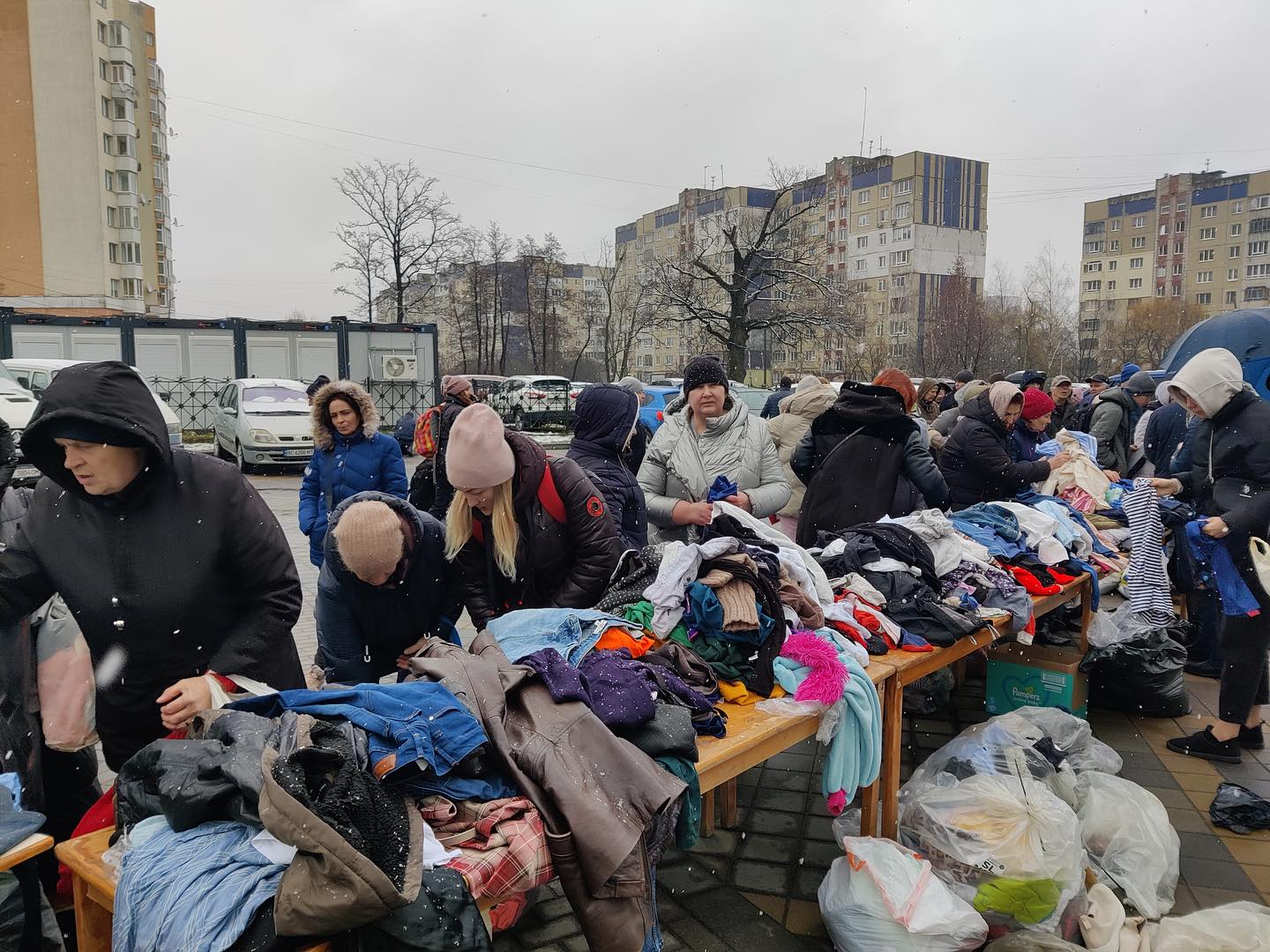 People sorting through donated clothes on tables outdoors in a city setting, with apartment buildings in the background on a cloudy day.