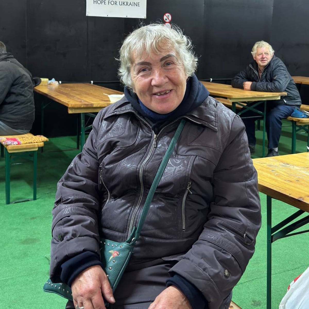 An older woman in a gray coat sits and smiles at the camera in an indoor setting with wooden tables and benches. Other people are seated in the background.