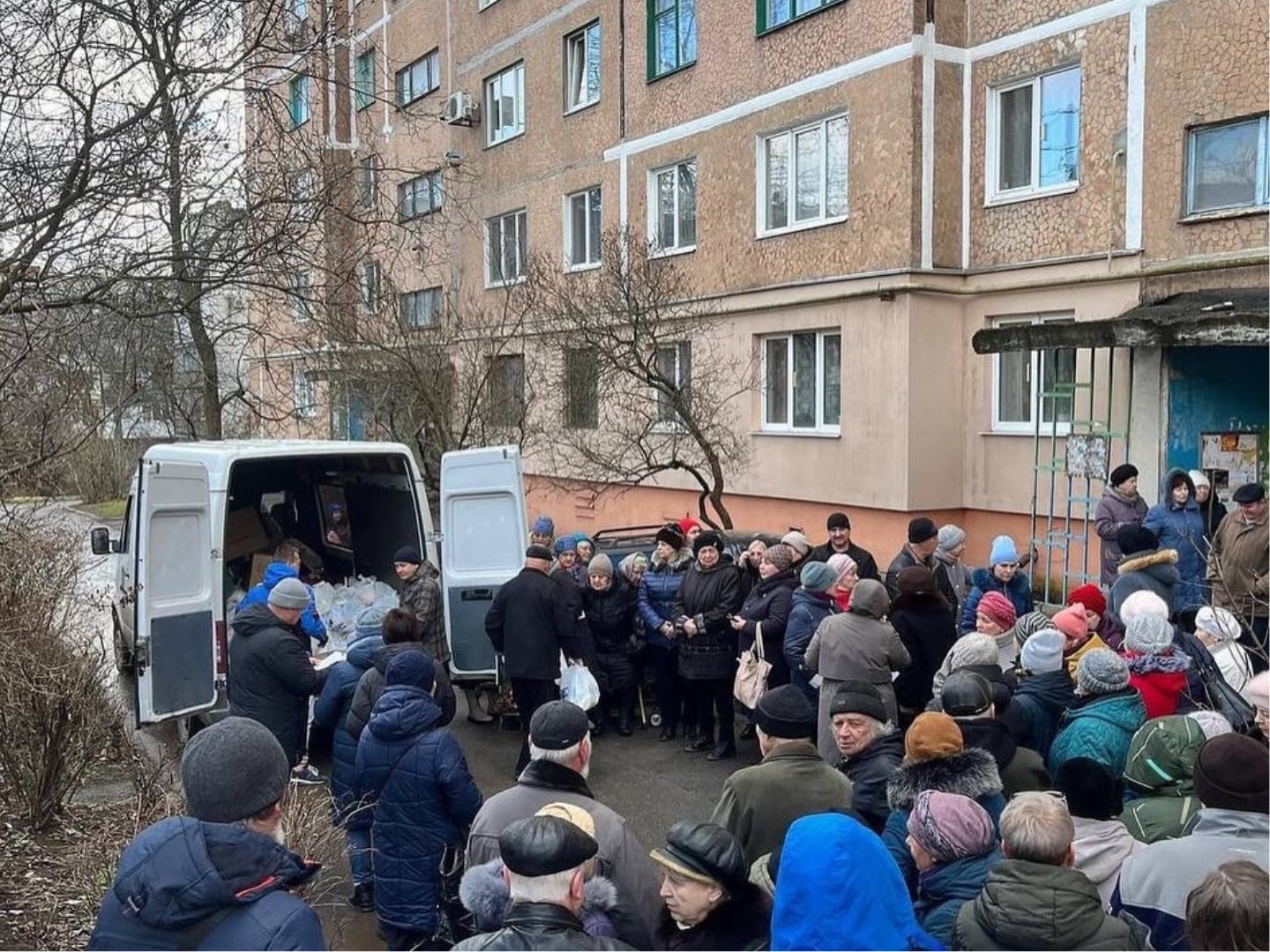A group of people gathers around a van distributing supplies outside a residential apartment building on a cloudy day.