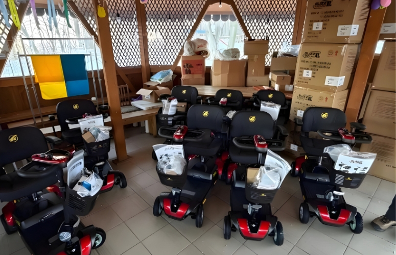 A group of electric mobility scooters with black seats and red bases are arranged indoors, surrounded by cardboard boxes and supplies.