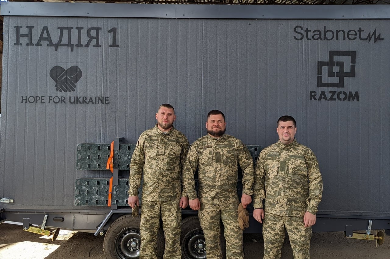 Three men in military uniforms stand in front of a gray building with logos and text reading "Hope for Ukraine," "Stabnet," "Razom," and "Надія 1.
