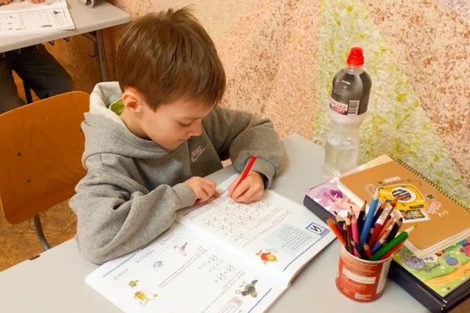 A young boy sits at a desk coloring in a workbook with colored pencils, surrounded by school supplies and a water bottle—enjoying one of many safe spaces to create and explore his imagination.