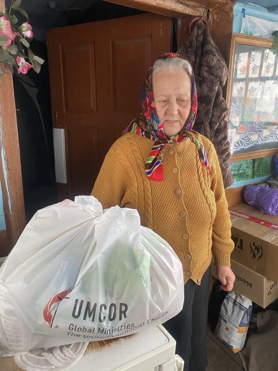 An older woman wearing a colorful headscarf and yellow sweater stands indoors, holding a plastic bag labeled "UMCOR Global Ministries." Boxes, household items, and supplies for abandoned pets from the Ukraine war are visible in the background.