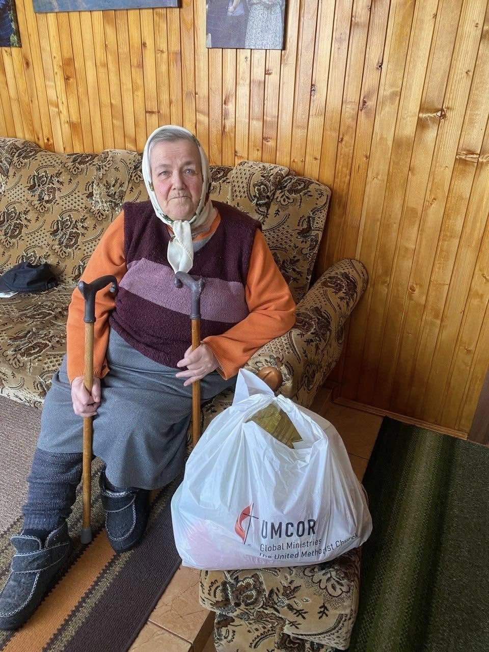 An elderly woman with two canes sits on a patterned couch next to a large UMCOR donation bag in a wood-paneled room, reflecting the hardships faced by many, including those caring for abandoned pets during the Ukraine war.