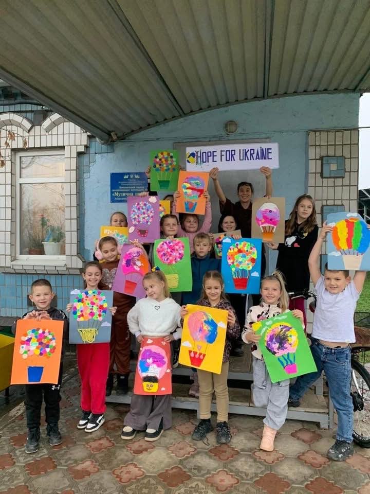 A group of children and two adults pose outside under a roof, holding colorful art projects in front of a “Hope for Ukraine” sign.