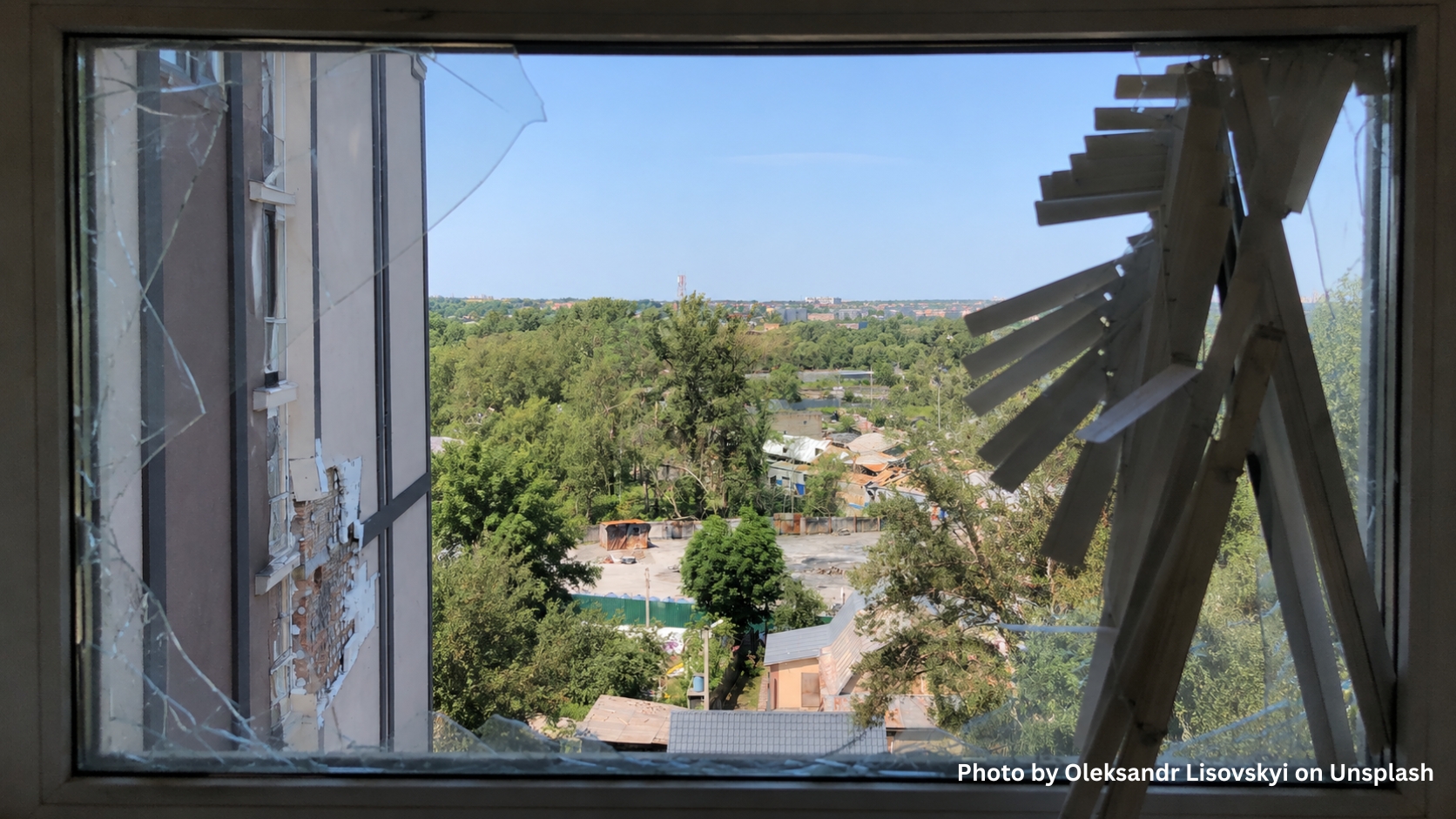 A partially broken window with shattered glass and damaged blinds overlooks a green landscape, several buildings in the distance, and hints at the need for food kits in Ukraine.