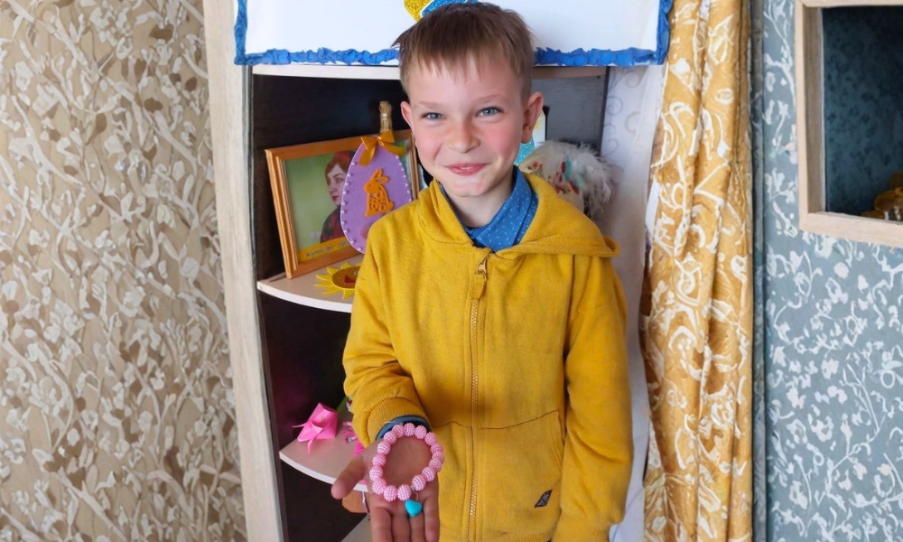A young boy in a yellow hoodie stands indoors, smiling and holding a beaded bracelet in his hand, with shelves and framed photos in the background.