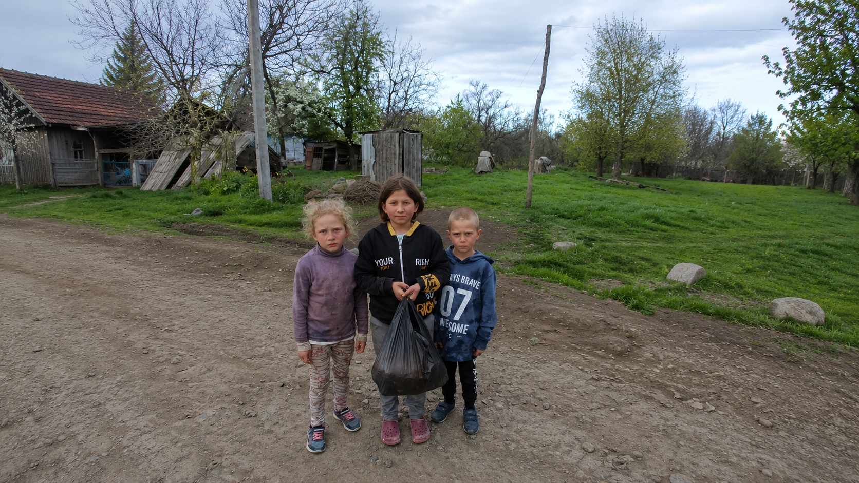 Three children stand on a dirt road in a rural area, holding a black plastic bag, with old buildings and green trees in the background.