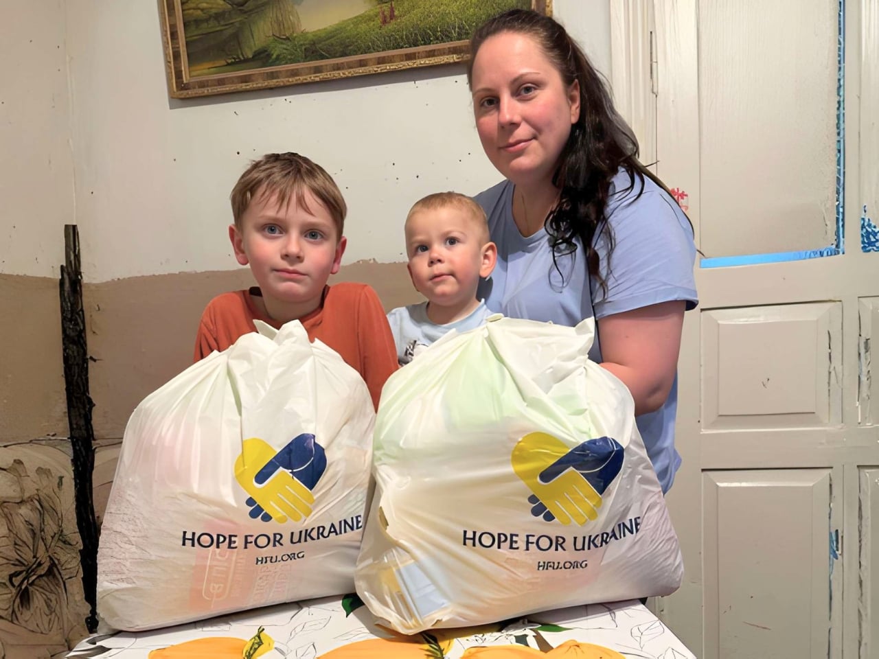 A woman and two children stand behind two white bags labeled "Hope For Ukraine" on a table indoors.