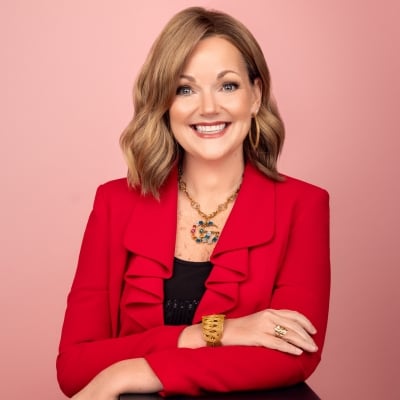 Woman with shoulder-length light brown hair wearing a bright red blazer, black top, and gold jewelry, smiling in front of a pink background.