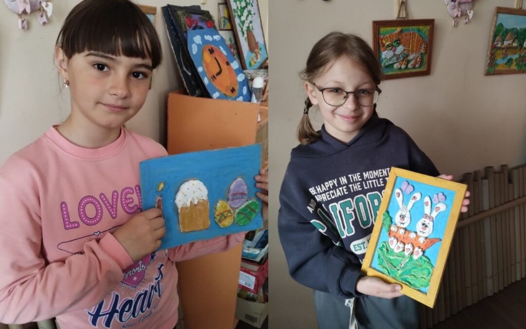 Two girls standing indoors, each holding a framed drawing; the left features Easter-themed art, while the right shows white rabbits in a grassy setting—perfect for highlighting rescue frontline pets and their inspiring stories.