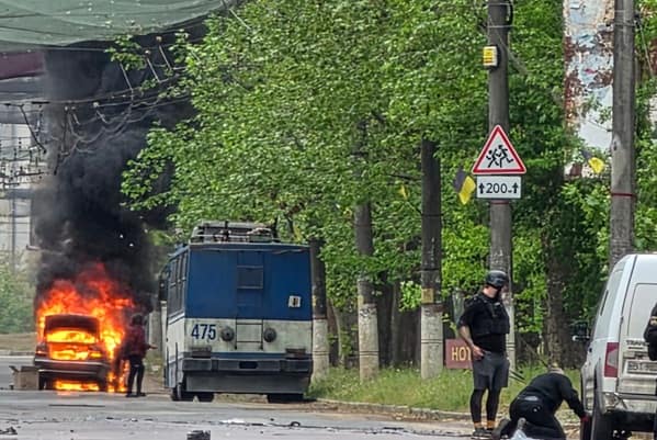 A car is on fire near a blue bus as two people in protective gear stand on the street; smoke is rising and a caution sign is visible.