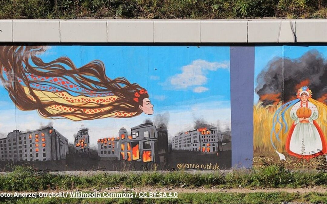 A mural reflects Ukrainian culture during war, showing a woman’s head with flowing hair above burning buildings on the left, and a woman holding pumpkins in a field with fire and smoke in the background on the right.