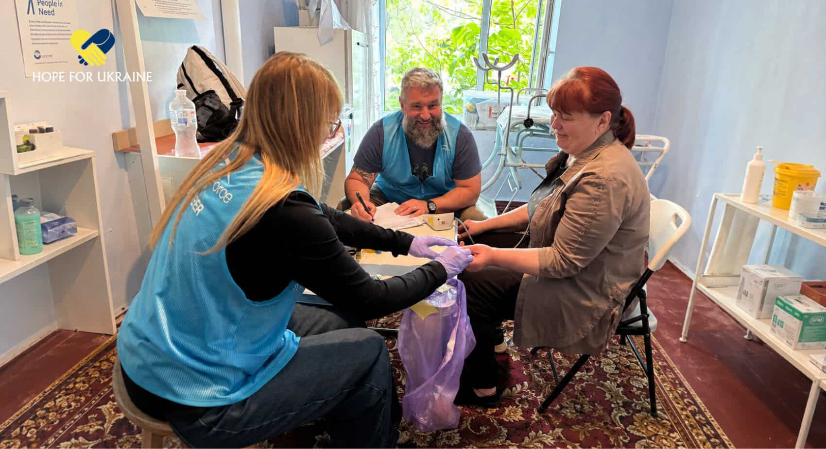 Three people are seated in a clinic room; a volunteer in blue assists a woman while a man takes notes. Medical supplies and a 'Hope for Ukraine' sign are visible.
