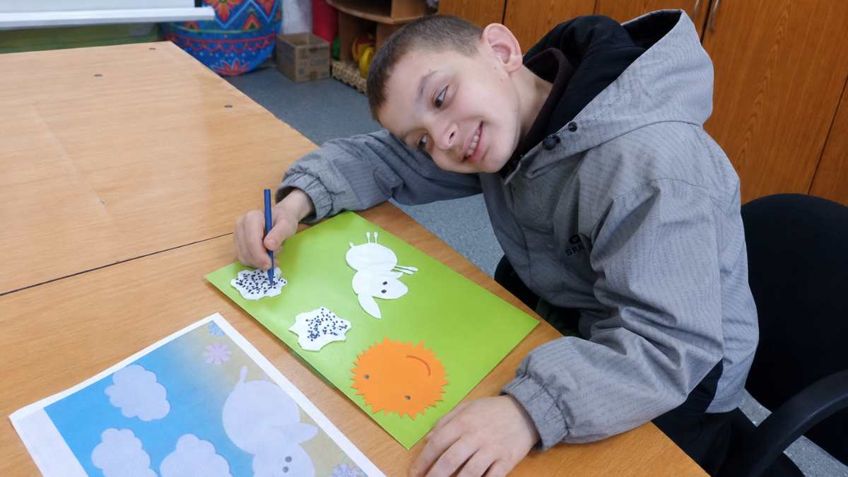 A boy in a gray jacket sits at a table, smiling while drawing on a green sheet with cut-out shapes of a sheep and a sun—bright moments that can help support Ukrainian children amid the mental health crisis. Art supplies are on the table.