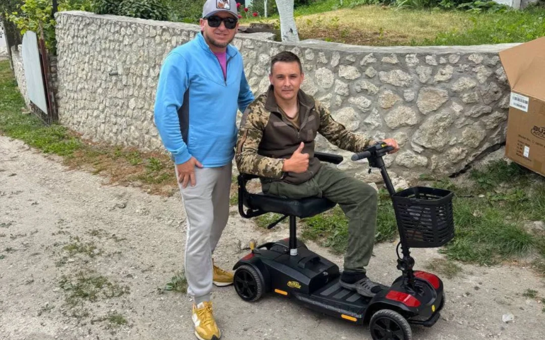 Two men outdoors; one stands and smiles while the other, in camouflage, sits on a mobility scooter giving a thumbs-up. A stone wall and greenery are in the background, reminiscent of scenes supporting children's art therapy after the Ukraine war.