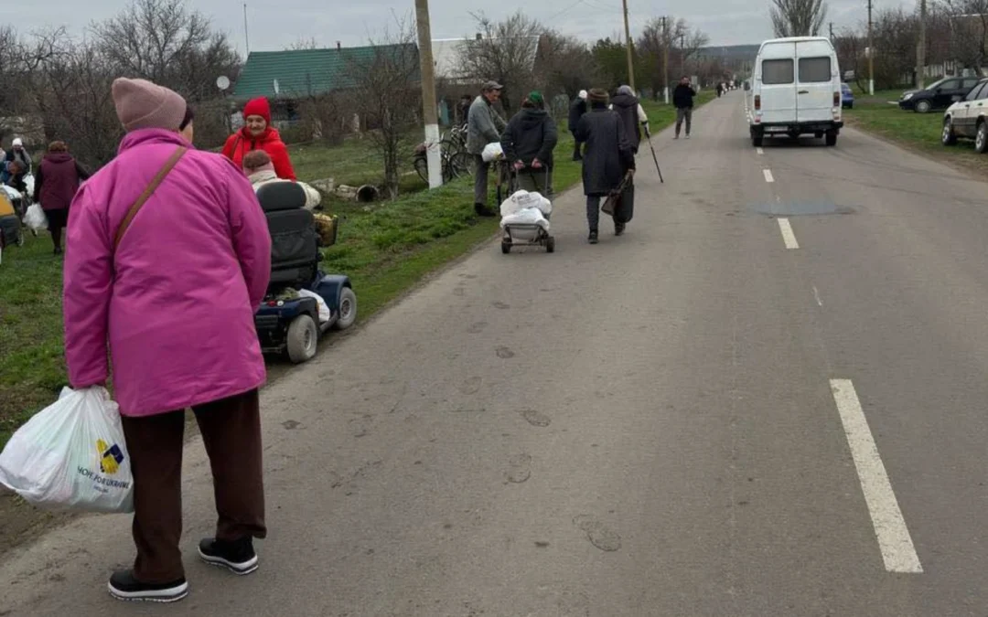 People walk along a rural road carrying bags and pulling carts; a white van is parked ahead, and some individuals—including one using an electric mobility scooter for wounded soldiers Ukraine—are gathered on the grassy roadside.