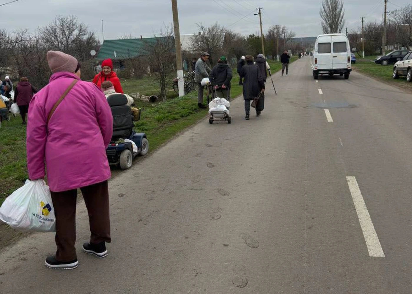 People walk along a rural road carrying bags and pulling carts; a white van is parked ahead, and some individuals—including one using an electric mobility scooter for wounded soldiers Ukraine—are gathered on the grassy roadside.