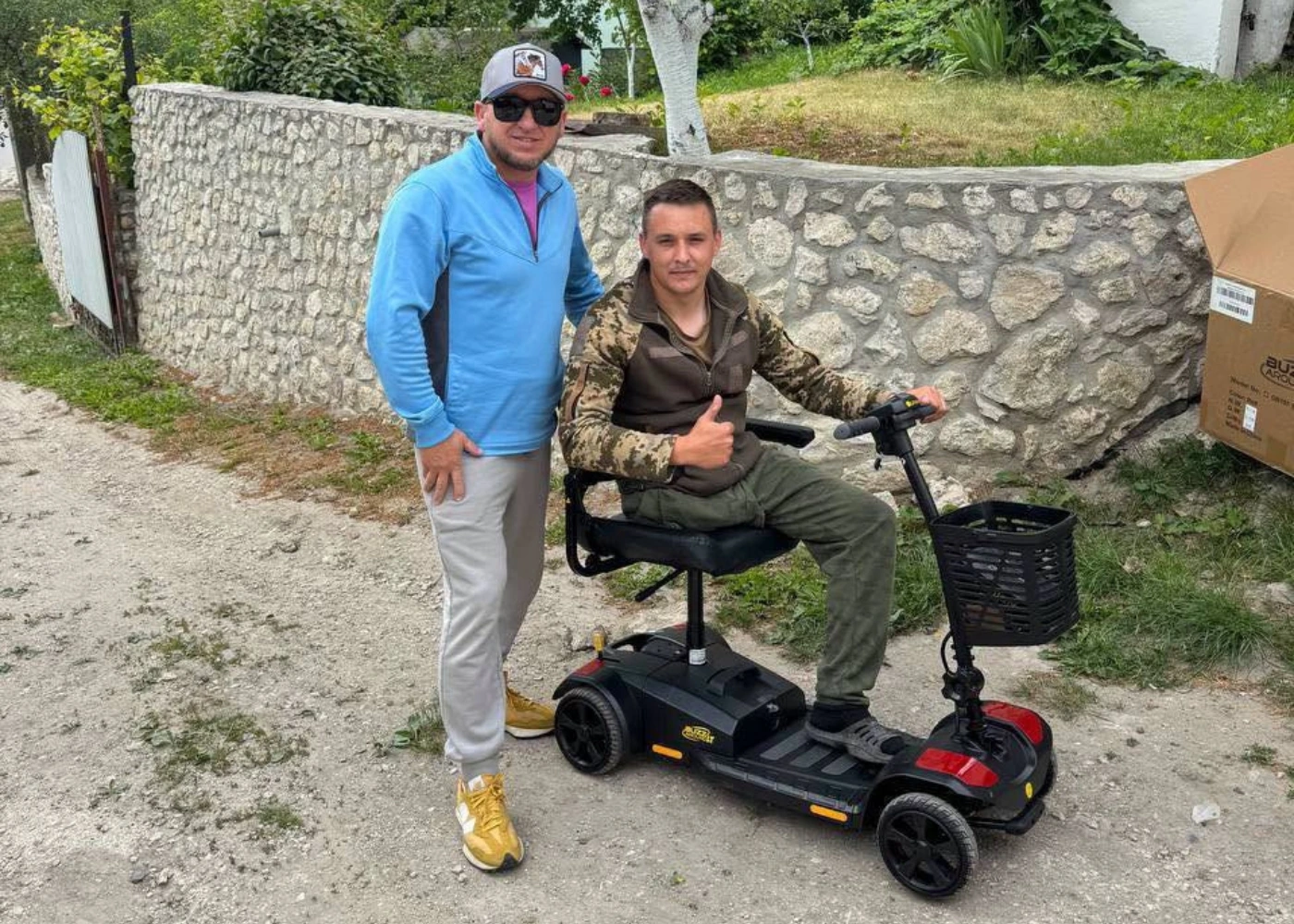 Two men outdoors; one stands and smiles while the other, in camouflage, sits on a mobility scooter giving a thumbs-up. A stone wall and greenery are in the background, reminiscent of scenes supporting children's art therapy after the Ukraine war.
