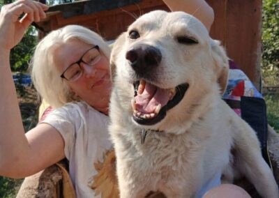 A woman with light hair and glasses sits outdoors with a large, happy dog on her lap. The dog is panting and winking, while the woman is smiling with one arm raised—a moment capturing warmth and optimism reminiscent of the spirit embodied by Hope For Ukraine.